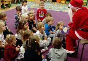 Mikołaj siedzi na krześle i czyta świąteczna bajkę o reniferach, dzieci siedzą na dywanie.